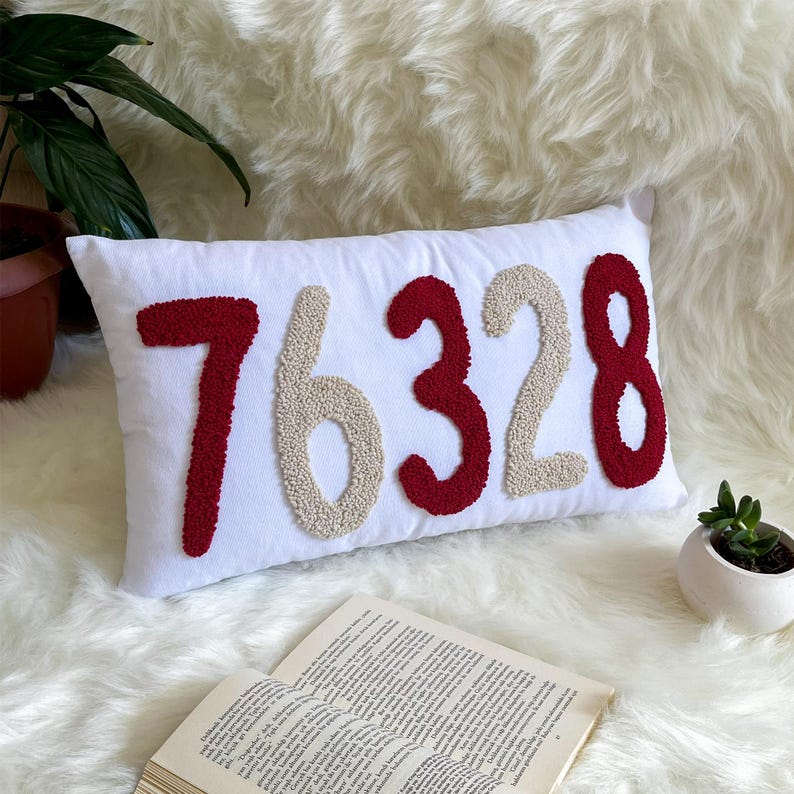 May include: White rectangular cushion featuring the number "76328" embroidered in red and beige. The cushion is on a white fluffy surface, with a plant and a book in the background.