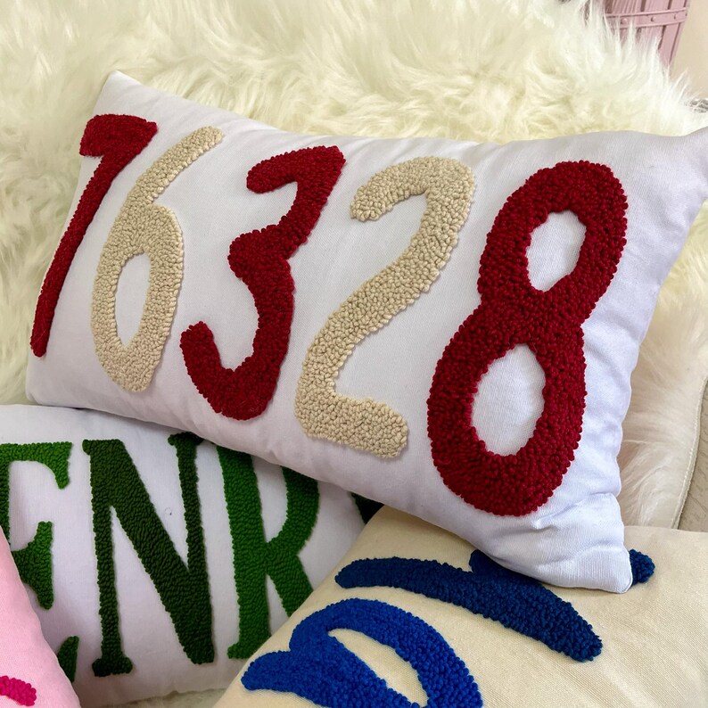May include: White rectangular cushion featuring the numbers 16328 in red and beige textured fabric. The cushion is on a white fluffy surface. Other cushions with letters and numbers are visible.