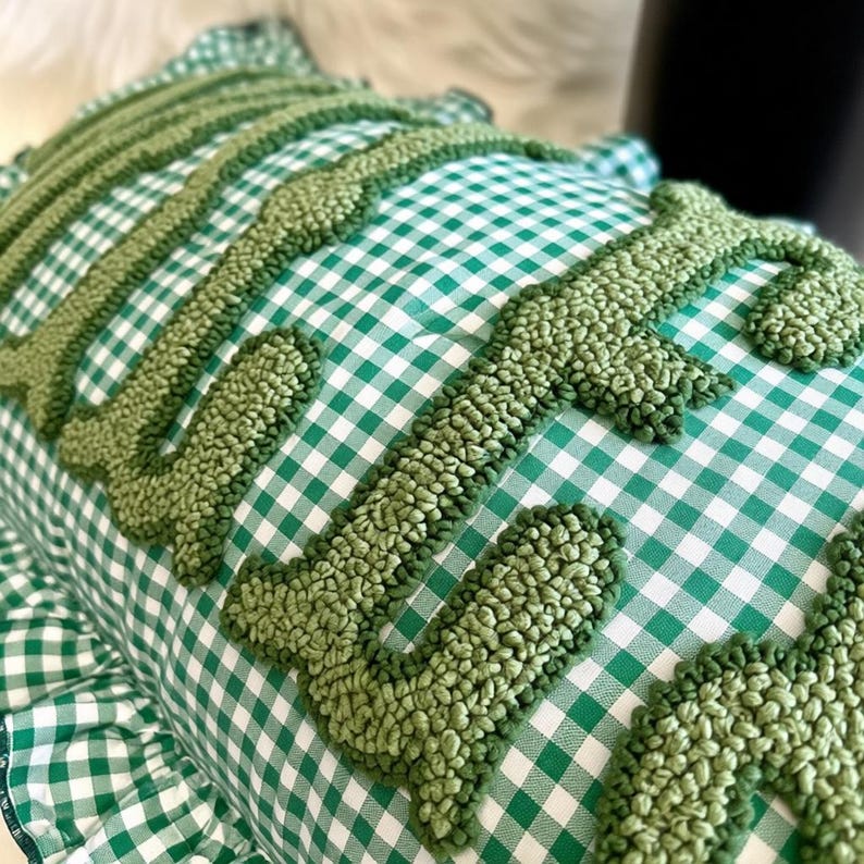 May include: A cushion with a green and white gingham pattern, featuring the word "LOVE" embroidered in a textured, light green yarn. The cushion has a ruffled edge and is a decorative home accent.
