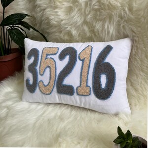 May include: White rectangular cushion featuring the number "35216" embroidered in grey, beige, and blue. The cushion is set on a fluffy, white surface, with green plants in the background.