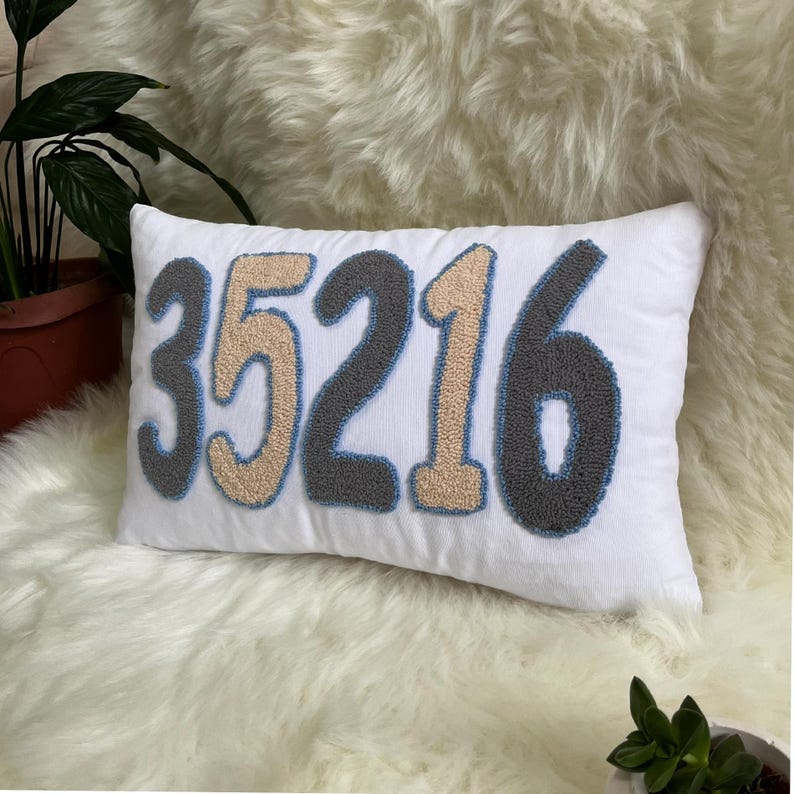 May include: White rectangular cushion featuring the number "35216" embroidered in grey, beige, and blue. The cushion is set on a fluffy, white surface, with green plants in the background.