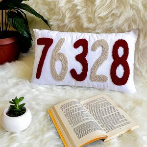 May include: A white rectangular cushion embroidered with the numbers "76328" in burgundy and beige. The cushion rests on a fluffy white surface, with a small potted plant and an open book in view.