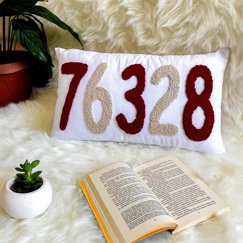 May include: A white rectangular cushion embroidered with the numbers "76328" in burgundy and beige. The cushion rests on a fluffy white surface, with a small potted plant and an open book in view.