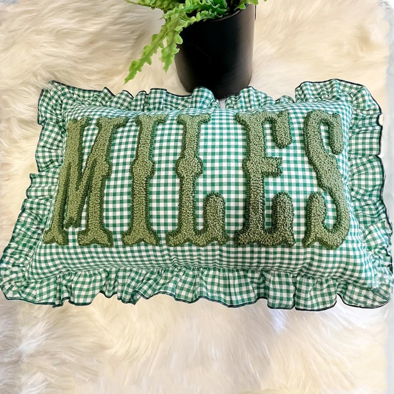 May include: A rectangular cushion with a green and white gingham pattern, featuring the name "MILES" in a textured, darker green embroidery. The cushion has a ruffled border. It is placed on a white, fluffy surface.