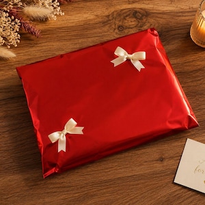 May include: A wrapped gift in shiny red paper, adorned with two small ivory bows. A lit candle in a glass holder and a card that reads "A gift for you" are on a wooden surface.