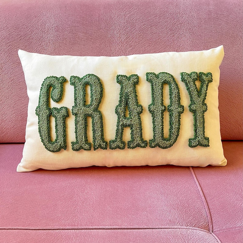 Puede incluir: Coj&iacute;n rectangular crema con el nombre "GRADY" bordado en letras verdes y texturizadas. El coj&iacute;n est&aacute; colocado sobre un sof&aacute; rosa. El dise&ntilde;o es simple y elegante, con un claro contraste entre los colores.