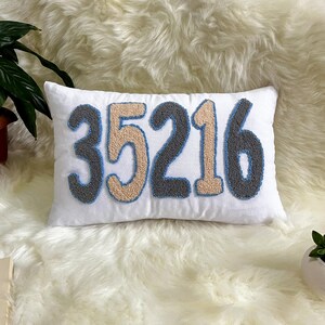 May include: White rectangular cushion featuring the embroidered number "35216" in grey, tan, and blue. The cushion is on a white fluffy surface, with a plant in the background.