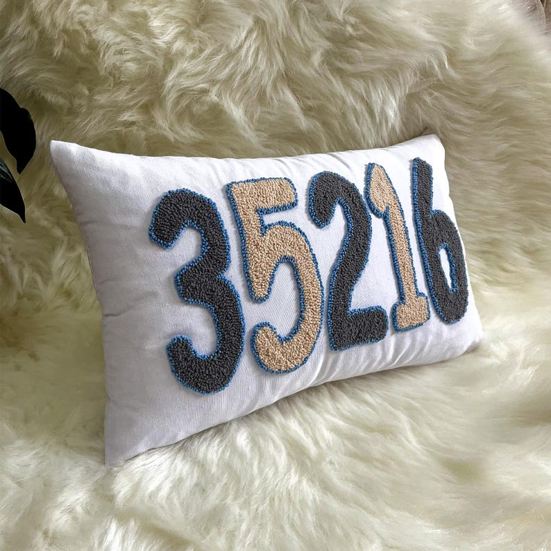 May include: White rectangular cushion featuring the numbers 35216 in a textured embroidery style. The numbers are in grey, beige, and black with blue outlines. The cushion rests on a white, fluffy surface.