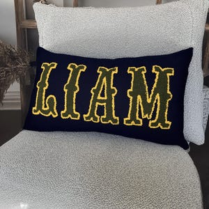 Personalized boho style name pillow with raised yellow punch needle letters on navy fabric, placed on a textured chair for modern home accents.