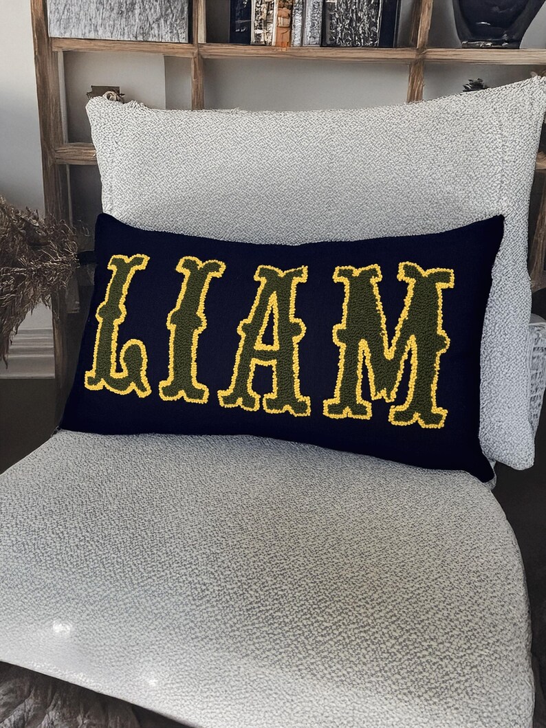 Personalized boho style name pillow with raised yellow punch needle letters on navy fabric, placed on a textured chair for modern home accents.