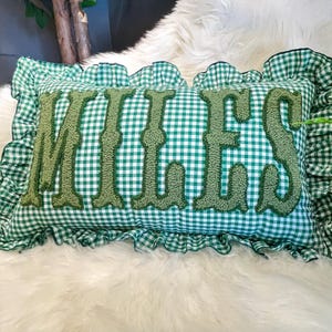 May include: A rectangular cushion featuring a green and white gingham design. The word "MILES" is embroidered in a textured green font across the centre. The cushion has a ruffled edge made from the same gingham fabric.