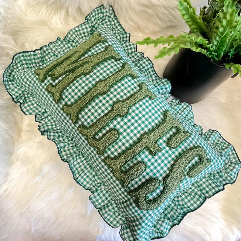 May include: A rectangular cushion with a green and white gingham pattern. The cushion has a ruffled edge and the word "Wifey" in a textured, green font across the centre. A fern in a black pot is in the background.