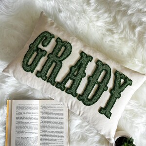 Puede incluir: Un coj&iacute;n rectangular blanco roto con el nombre "GRADY" bordado en una fuente verde oscuro texturizada. El coj&iacute;n est&aacute; colocado sobre una superficie blanca y esponjosa, con un libro abierto visible en primer plano.