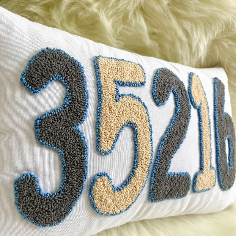 May include: White rectangular cushion featuring the embroidered numbers "35216" in grey, beige, and blue. The numbers have a textured, raised design. A decorative home accessory.