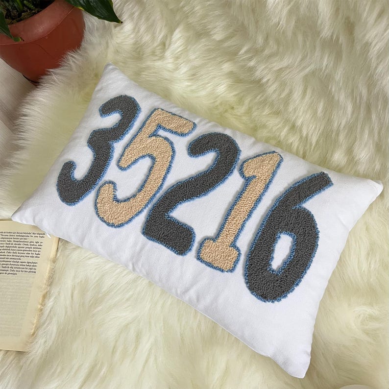 May include: White rectangular cushion embroidered with the number 35216 in grey, beige, and blue. The cushion rests on a white fluffy surface. A potted plant is visible in the background.