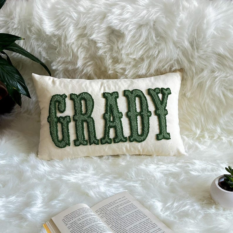 Puede incluir: Un coj&iacute;n rectangular blanco roto con el nombre "GRADY" bordado en verde oscuro, con una fuente texturizada. El coj&iacute;n est&aacute; colocado sobre una superficie de piel sint&eacute;tica blanca y esponjosa. Tambi&eacute;n se ven una planta en maceta y un libro abierto.