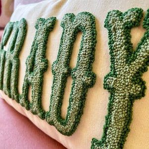Puede incluir: Coj&iacute;n rectangular color crema con la palabra "READY" bordada en hilo verde texturizado. Las letras son grandes y estilizadas, con un efecto tridimensional en relieve. El coj&iacute;n est&aacute; sobre una superficie rosa.