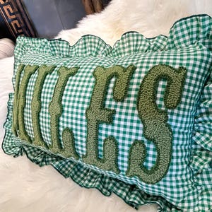 May include: A rectangular cushion with a green and white gingham pattern. The word "Kisses" is embroidered in a textured, olive green font across the centre. The cushion has a ruffled edge in the same gingham fabric.