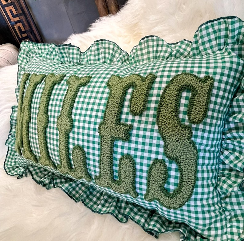 May include: A rectangular cushion with a green and white gingham pattern. The word "Kisses" is embroidered in a textured, olive green font across the centre. The cushion has a ruffled edge in the same gingham fabric.