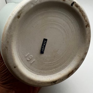 May include: The bottom of a light blue ceramic vase. The base is off-white with a dark ring around the edge. A small black label reads "JAPAN". The number "191" is stamped on the base.
