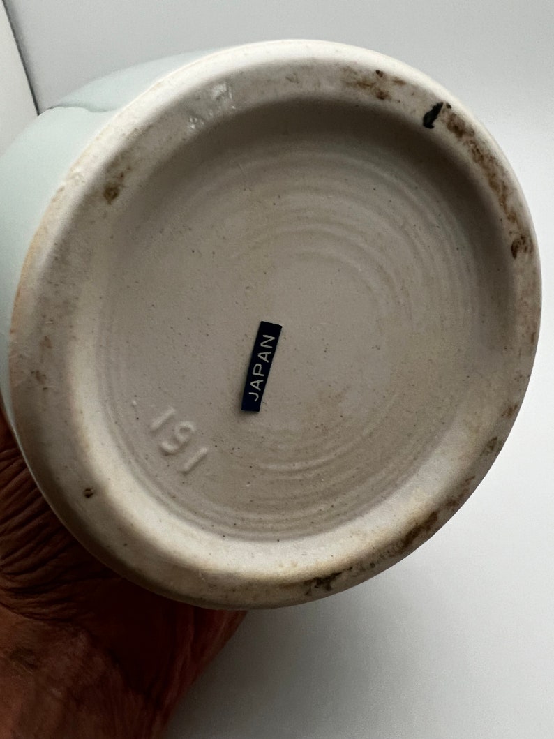 May include: The bottom of a light blue ceramic vase. The base is off-white with a dark ring around the edge. A small black label reads "JAPAN". The number "191" is stamped on the base.