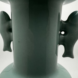 May include: A close-up of a celadon-colored ceramic vase with two fish-shaped handles. The vase has a wide base and a flared top. The handles are detailed with fins and eye-like features.