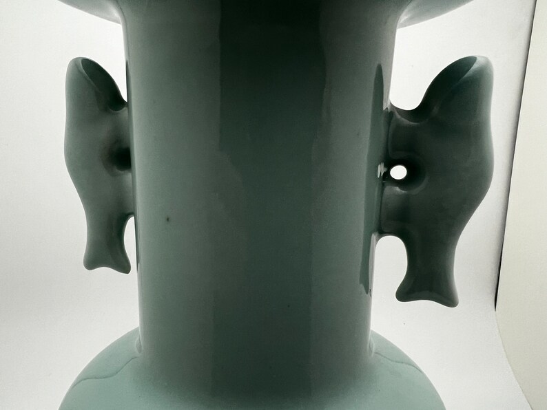 May include: A close-up of a celadon-colored ceramic vase with two fish-shaped handles. The vase has a wide base and a flared top. The handles are detailed with fins and eye-like features.