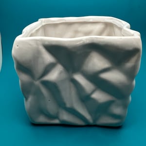 May include: A white ceramic planter with a textured, crumpled paper bag design. The square-shaped container has a wide opening and sits on a teal surface. The planter is suitable for small plants or decorative use.