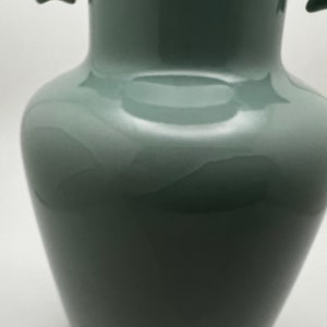 May include: A tall, glossy, sage green vase with a bulbous body and a narrow neck. The vase has two small, decorative handles near the top. The smooth surface reflects light, highlighting its elegant shape. This decorative vase is suitable for home decor.