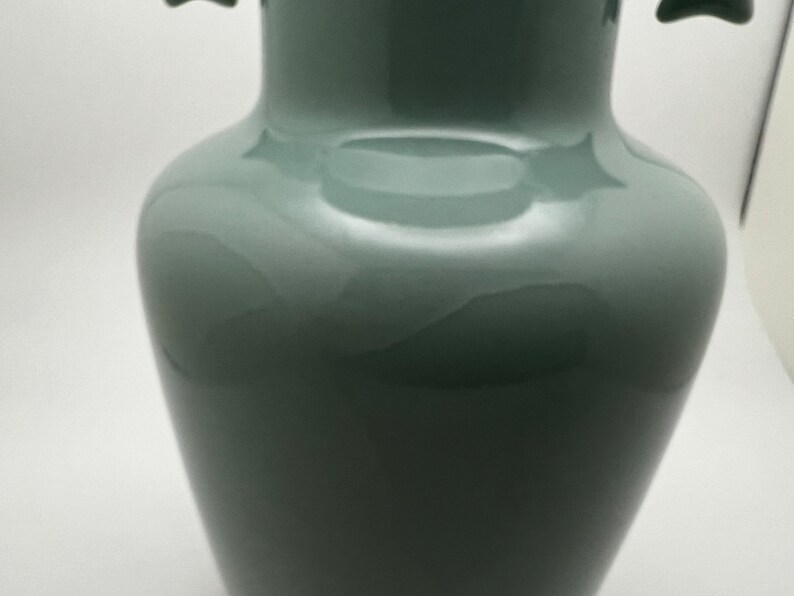 May include: A tall, glossy, sage green vase with a bulbous body and a narrow neck. The vase has two small, decorative handles near the top. The smooth surface reflects light, highlighting its elegant shape. This decorative vase is suitable for home decor.