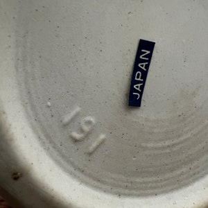 May include: Close-up of the bottom of a ceramic object. The object is off-white with a speckled texture. A small, dark blue label with the word "JAPAN" in white is affixed to the surface. The number "191" is stamped into the ceramic.