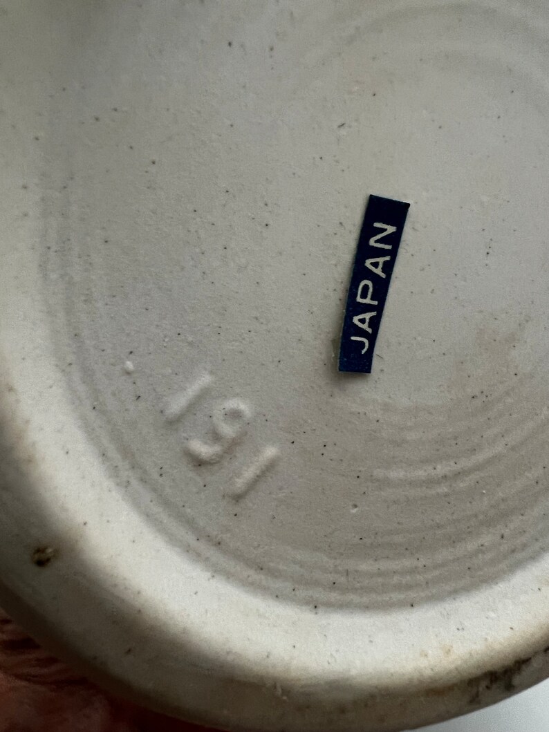 May include: Close-up of the bottom of a ceramic object. The object is off-white with a speckled texture. A small, dark blue label with the word "JAPAN" in white is affixed to the surface. The number "191" is stamped into the ceramic.