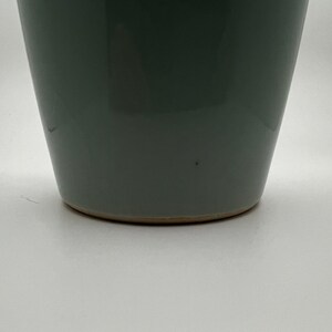 May include: A gray-green ceramic vase with a slightly tapered shape. The vase has a smooth, glossy finish and a light brown base. The design is simple and modern, suitable for various home decor styles.