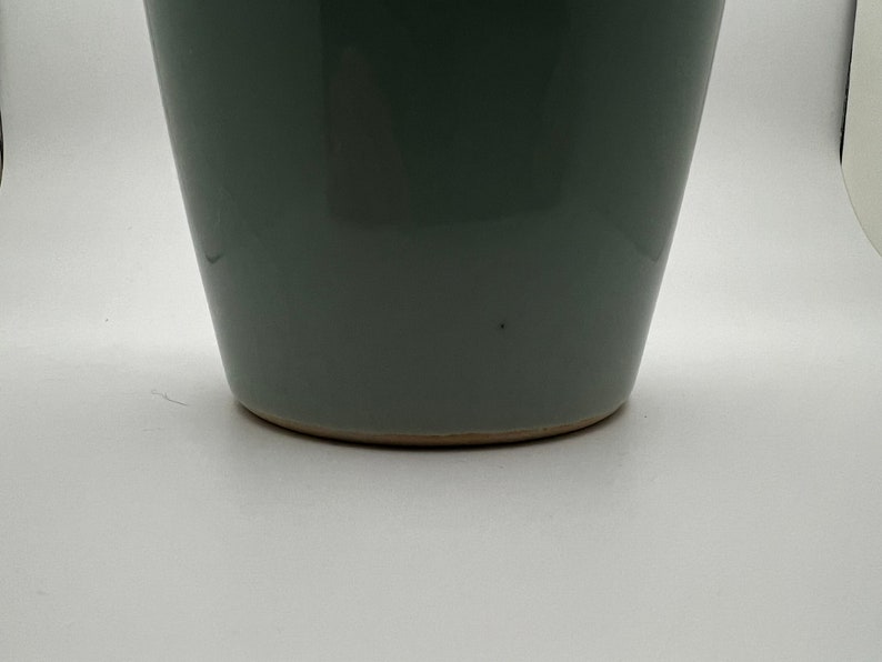 May include: A gray-green ceramic vase with a slightly tapered shape. The vase has a smooth, glossy finish and a light brown base. The design is simple and modern, suitable for various home decor styles.