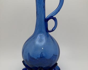 May include: A cobalt blue glass pitcher with a rounded body and a slender, curved handle. The pitcher has a flared spout and sits on a base with six rounded feet. The glass has a slight swirl pattern.