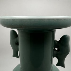 May include: A celadon-colored ceramic vase with a wide, flat top and a cylindrical body. The vase features two decorative, stylized handles on the sides. The overall design is simple and elegant, with a smooth, glossy finish.