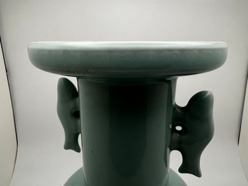 May include: A celadon-colored ceramic vase with a wide, flat top and a cylindrical body. The vase features two decorative, stylized handles on the sides. The overall design is simple and elegant, with a smooth, glossy finish.