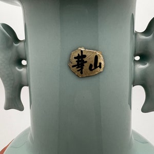 May include: A celadon-colored ceramic vase with two side handles. The vase has a flared top and a round base. A small, gold-colored label with black Chinese characters is affixed to the front of the vase. The vase is set against a white background.