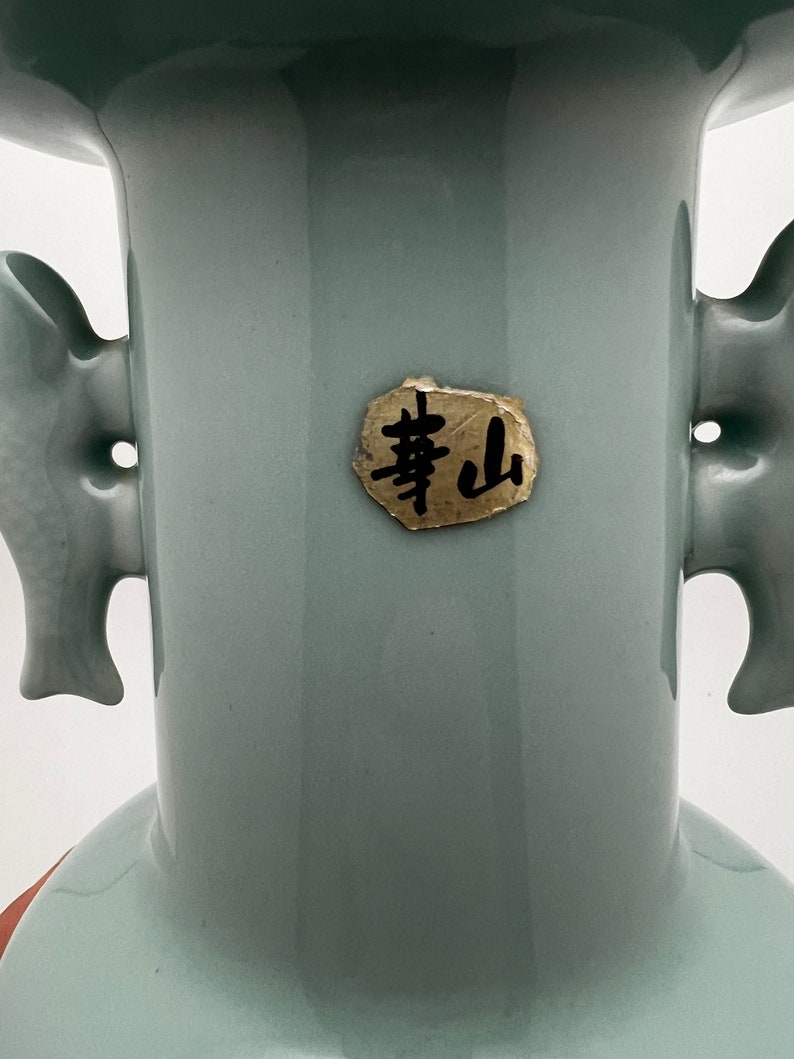 May include: A celadon-colored ceramic vase with two side handles. The vase has a flared top and a round base. A small, gold-colored label with black Chinese characters is affixed to the front of the vase. The vase is set against a white background.