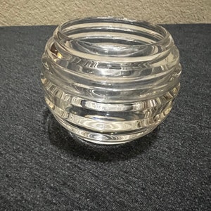 May include: A clear, round glass bowl with a ribbed design. The bowl has a wide opening and sits on a dark gray surface. The glass reflects light, highlighting the horizontal ridges that encircle the bowl.