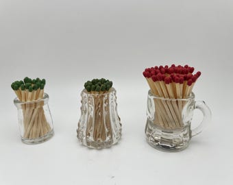May include: Three clear glass containers filled with matches. One has green tips, another has green tips, and the third has red tips. The containers vary in shape, including a small jar and a mug.