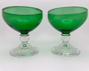 May include: Two green champagne coupe glasses. The bowls are a vibrant emerald green, and the stems are clear glass with a decorative base. The base has a textured, patterned design. The glasses are set against a white background.