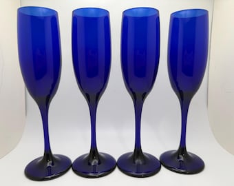 May include: Four cobalt blue champagne flutes. The glasses have a tall, slender design with a dark base and stem. The bowls of the flutes are a vibrant blue, and the stems and bases are a darker shade of blue.