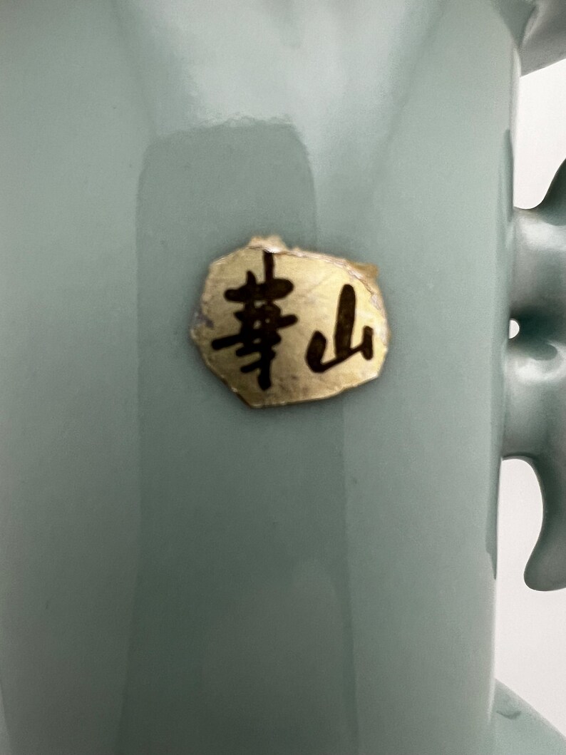 May include: A close-up of a pale blue ceramic vase with a handle. A gold-colored sticker with black Japanese characters is affixed to the vase. The vase has a smooth, glossy finish.