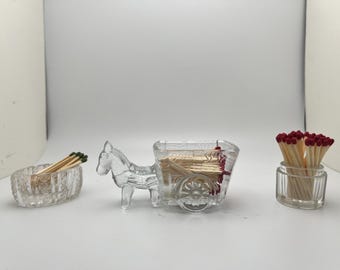 May include: Three clear glass match holders. One is a rectangular dish, another is a horse-drawn cart, and the third is a small round jar. All are filled with matches with red and green tips.