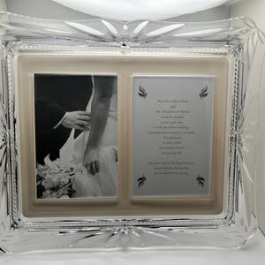 May include: A clear, rectangular crystal picture frame with a double opening. One side holds a black and white photo of a wedding, the other side has printed text. The frame has a decorative, starburst design on the corners.