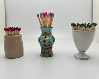 May include: Three decorative match holders filled with matches. A tan ceramic jar holds matches with red tips. A colorful, patterned vase contains matches with pink and blue tips. A white, footed holder has matches with green tips.