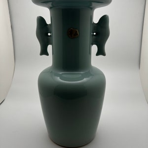 May include: A celadon-colored ceramic vase with a wide, flared rim and two fish-shaped handles. The vase has a rounded body and a small, circular seal near the center. The overall design is simple and elegant.