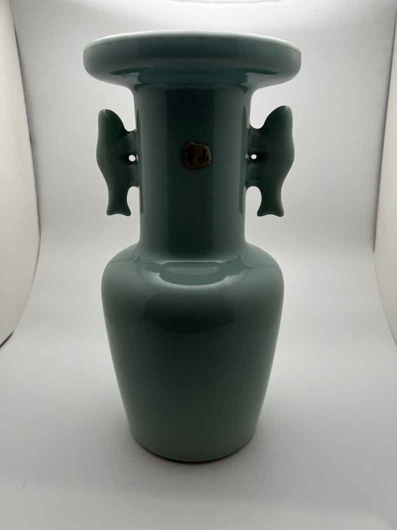 May include: A celadon-colored ceramic vase with a wide, flared rim and two fish-shaped handles. The vase has a rounded body and a small, circular seal near the center. The overall design is simple and elegant.