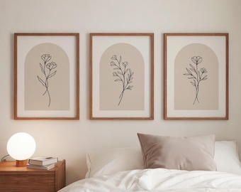 Puede incluir: Tres láminas botánicas enmarcadas. Cada lámina presenta un dibujo lineal negro de una flor sobre un fondo beige, enmarcado en un marco de madera marrón claro. Las láminas se exhiben sobre una cama con ropa de cama blanca y una almohada beige.